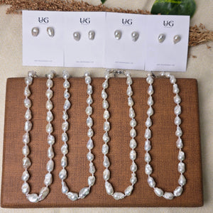 A set of white freshwater pearl jewelry, including a beaded necklace and matching earrings, displayed on a brown woven mat next to white card stands with size labels, set against a neutral beige fabric background with decorative green leaves.