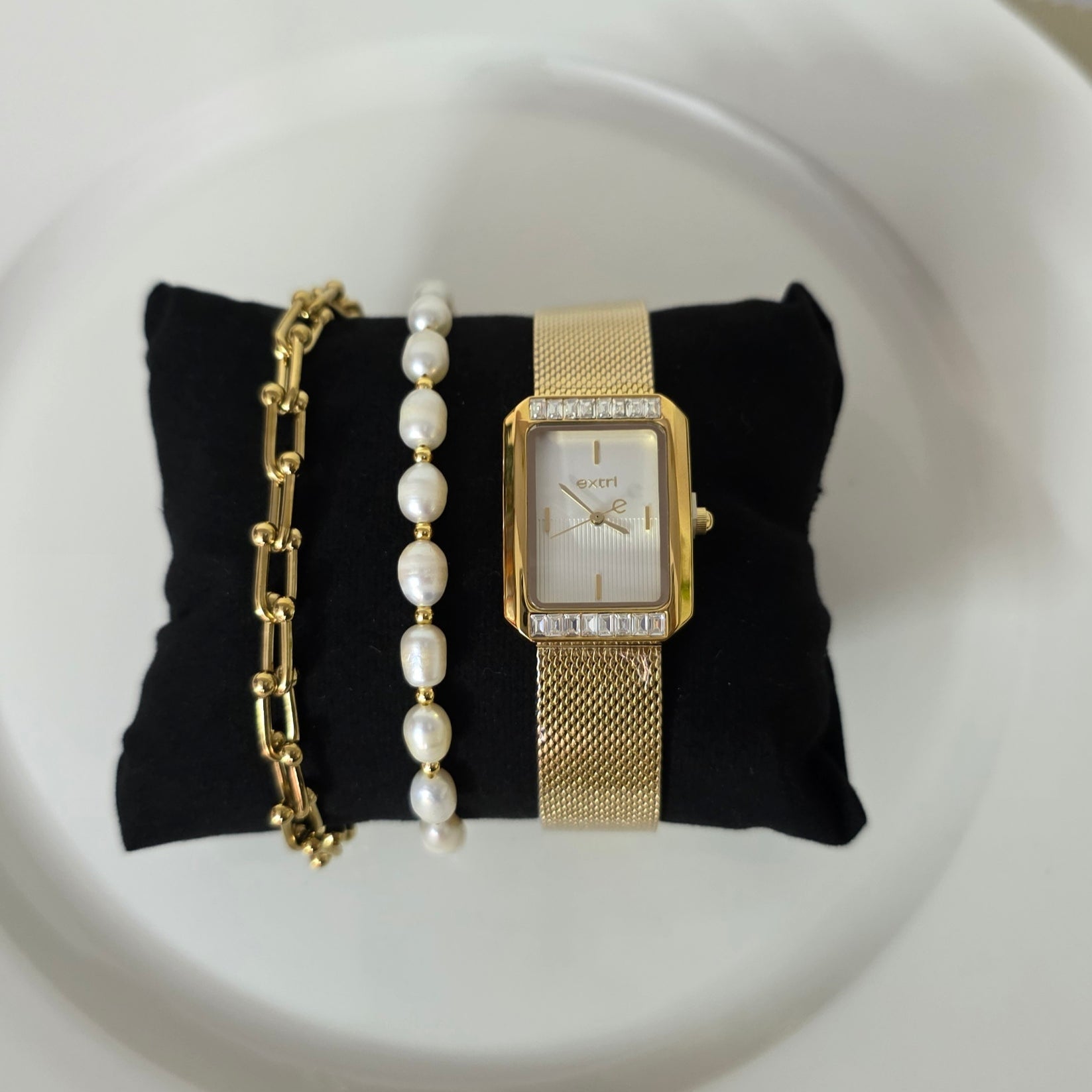 Gold watch, bracelet, and pearl necklace on a black fabric pad on a white plate.