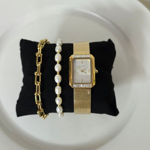 Gold watch, bracelet, and pearl necklace on a black fabric pad on a white plate.