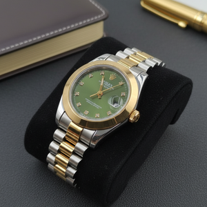 Luxury watch with green dial and gold accents on a black stand, with a book and pen in the background.