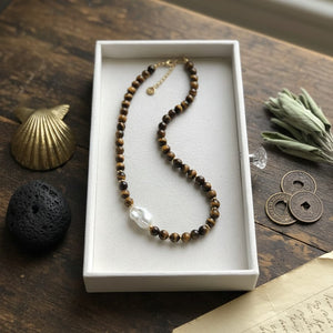 Necklace with brown and black beads in a white box on a wooden surface with a shell, black stone, and coins.