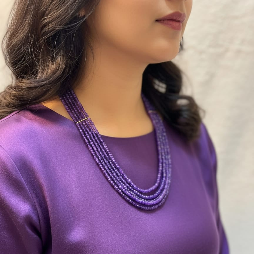 Woman wearing a purple top and multiple beaded necklaces against a neutral background