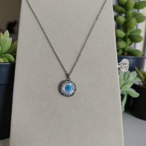 Necklace with a blue eye pendant on a beige card, plants in the background