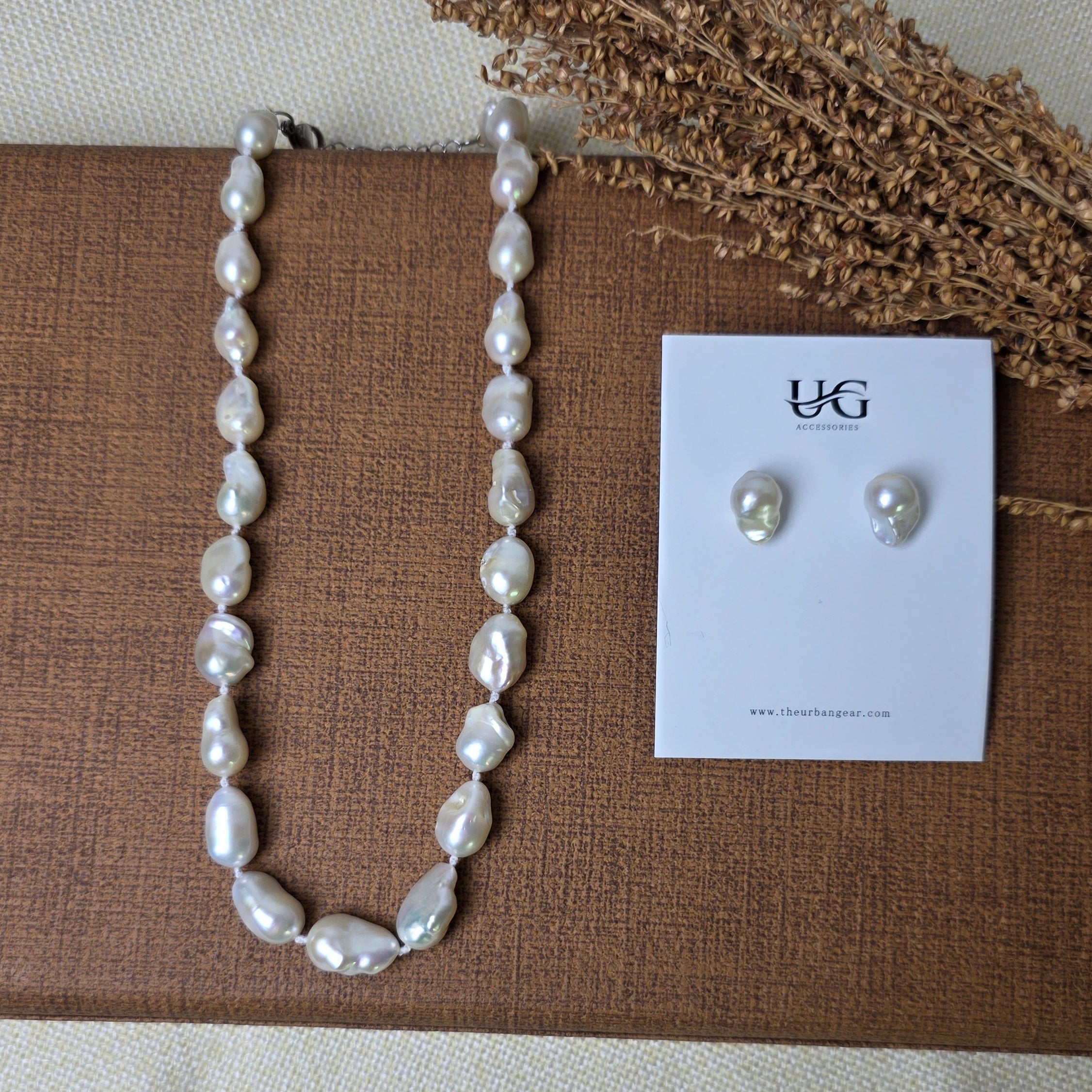 A pearl necklace and matching stud earrings displayed on a brown textured card, with dried grass stems on a light fabric background. The card shows the brand name 'YGG'.