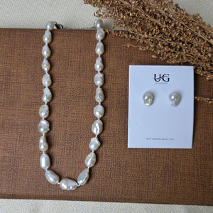 A pearl necklace and matching stud earrings displayed on a brown textured card, with dried grass stems on a light fabric background. The card shows the brand name 'YGG'.