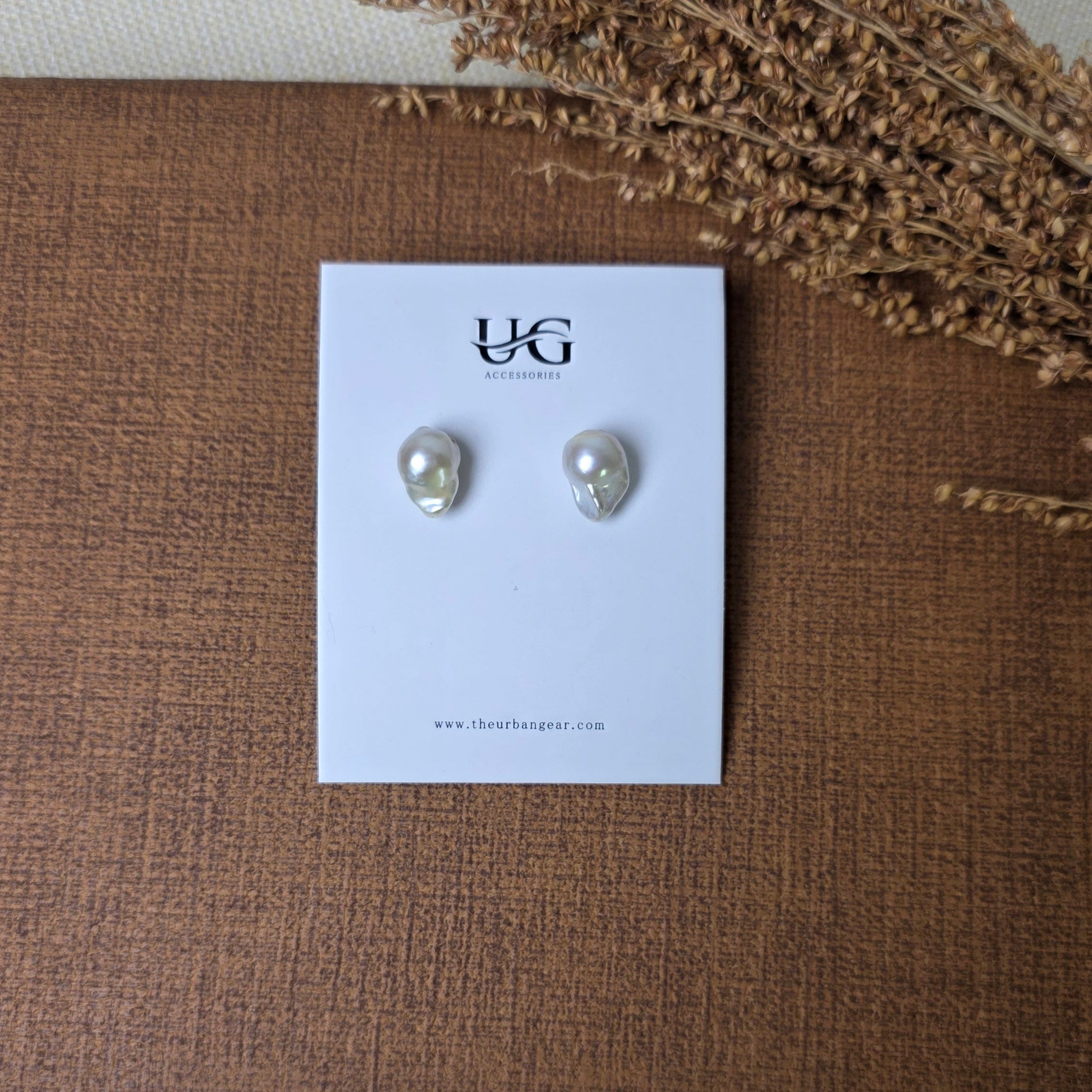 A pair of small silver stud earrings displayed on a white card, placed on a brown textured surface with dried floral stems and a light fabric background.