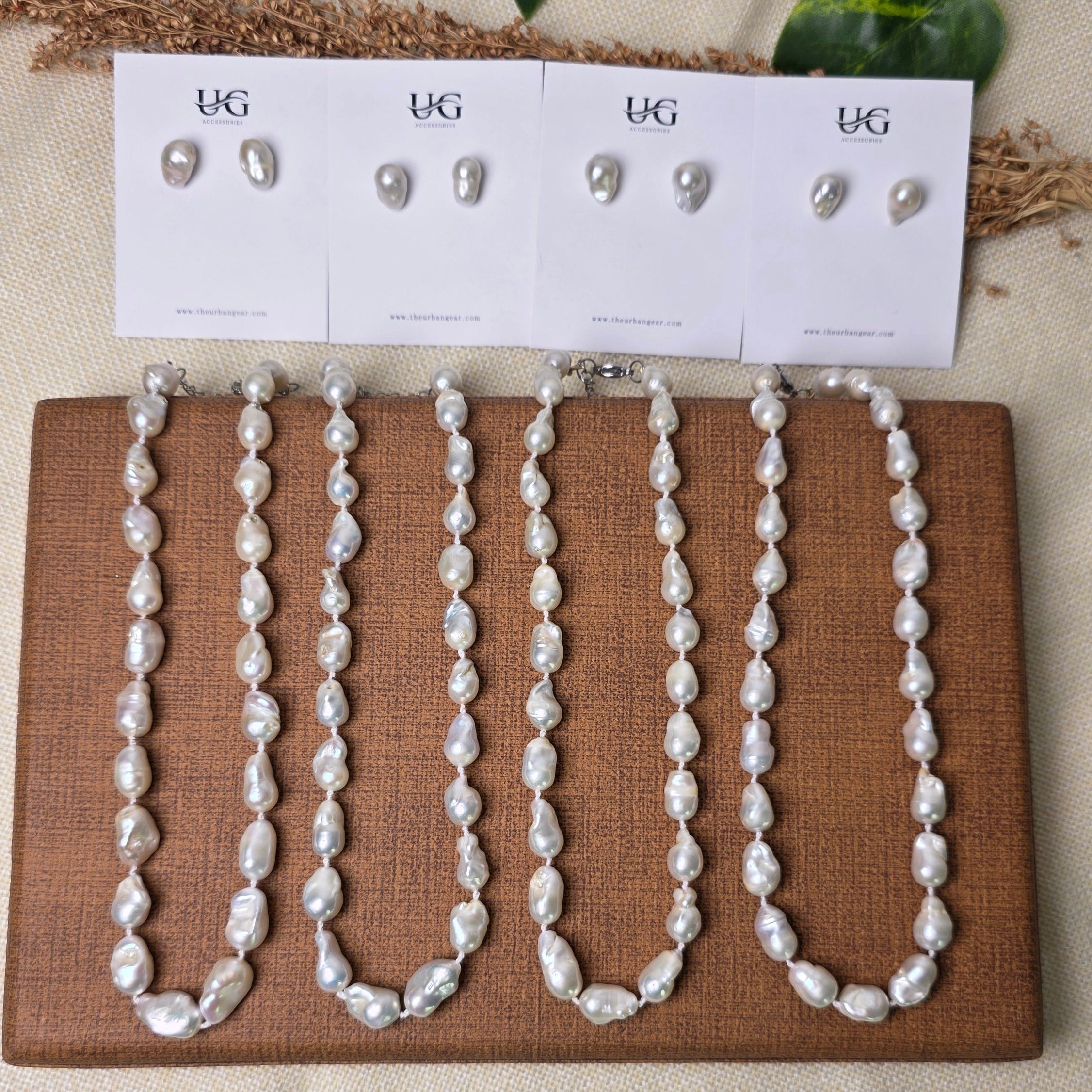 A set of white freshwater pearl jewelry, including a beaded necklace and matching earrings, displayed on a brown woven mat next to white card stands with size labels, set against a neutral beige fabric background with decorative green leaves.