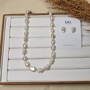 A pearl necklace and matching stud earrings displayed on a white jewelry organizer tray, with a small card labeled 'bk' and dried grass stems as styling props on a beige fabric background.