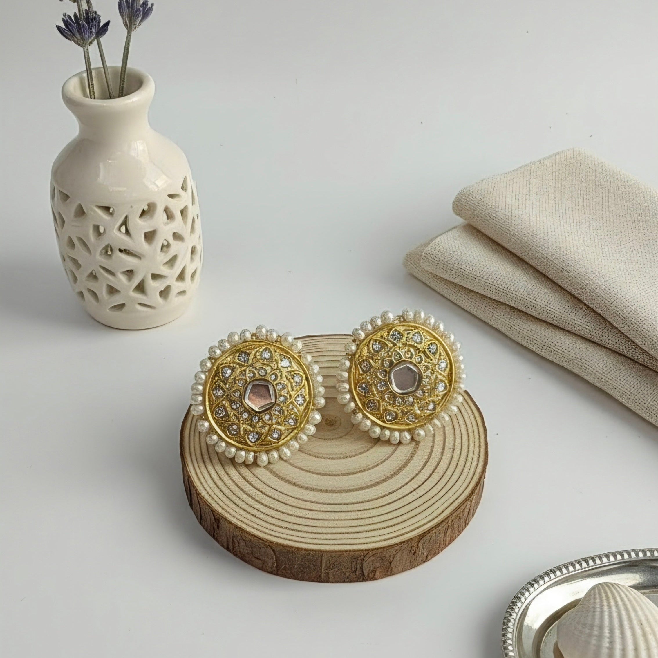 A pair of ornate gold and white beaded earrings displayed on a round wooden slice stand, styled on a light gray surface with a white vase of lavender, folded beige fabric, and a small silver dish.