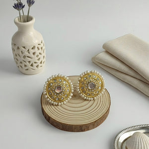 A pair of ornate gold and white beaded earrings displayed on a round wooden slice stand, styled on a light gray surface with a white vase of lavender, folded beige fabric, and a small silver dish.