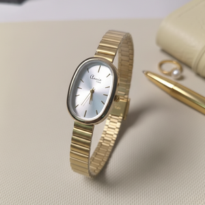 Gold watch on a light surface with a pen and jewelry box in the background
