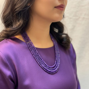 Woman wearing a purple top and multiple beaded necklaces against a neutral background